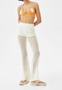 Szydełkowy pomarańczowy top na halter z białymi akcentami w postaci stokrotek, w połączeniu z białymi, prążkowanymi spodniami z dzianiny o rozszerzanym kroju i otwartej strukturze.