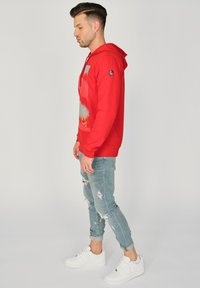 Rød hættetrøje med grafiktryk, snoretræk i huen og ribbede manchetter. Matchet med slidte lyseblå jeans og hvide sneakers.