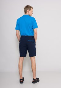 Blå polo tröja, marinblå shorts med en dragkedjeficka, avslappnat skor, visad från baksidan mot en neutral bakgrund.