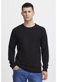 Pulover negru tricotat cu un model texturat, guler rotund și mâneci lungi, asociat cu blugi albaștri. Design simplu cu manșete cu rigle.