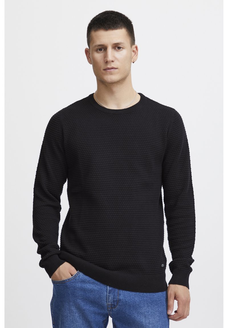 Pulover negru tricotat cu un model texturat, guler rotund și mâneci lungi, asociat cu blugi albaștri. Design simplu cu manșete cu rigle.