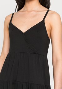 Czarna sukienka na cienkich ramiączkach, z dekoltem w kształcie litery V i marszczoną, warstwową spódnicą. Materiał wydaje się gładki i lekki, odpowiedni do codziennego noszenia.