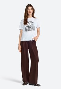 Weißes T-Shirt mit einer schwarzen, illustrierten Grafik einer Frau in einem Pullover. Kombiniert mit weiten braunen Hosen und dunklen Schuhen.