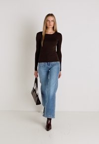 Hnedý top s dlhým rukávom v kombinácii so svetlomodrými širokými džínami. Modelka nesie tmavú kabelku a má na sebe hnedé špicaté topánky. Minimalistické pozadie.