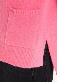 Maglione di lana rosa con taschino quadrato, orlo a coste e spacco laterale, abbinato a jeans neri di stoffa. Texture morbida visibile.