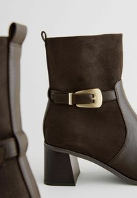 Bottines marron en cuir lisse et en daim, avec une accentuation en boucle dorée, un talon carré et une anse à l'arrière pour un enfilage facile.