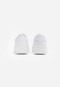 White sneakers with a textured leather upper, padded collar, and thick rubber sole. Subtle embossed logo detail on the heel.
