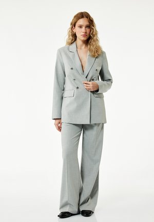 Vrouw met golvend blond haar die een lichtgrijze dubbelgeknoopte blazer en bijpassende wijde broek draagt, staand met één hand op de blazer.