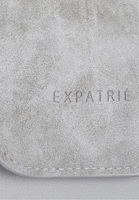Superficie in finta pelle grigia con una finitura texturizzata. Logo "EXPATRIÉ" in rilievo con un design sottile vicino alla parte inferiore, punto di cucitura lungo il bordo.