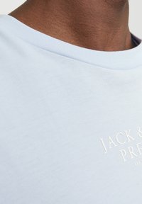 Světle modré bavlněné tričko s klasickým kulatým výstřihem. Má texturované bílé logo blízko hrudi. Hladká látka a uvolněný střih.