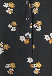 Svart stoff med blomstermønster i hvitt og gule nyanser, med runde svarte knapper langs fronten. Teksturert overflate.