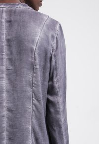 Giacca grigia, texture evidente con una superficie leggermente lucida, caratterizzata da cuciture visibili e un design semplice e strutturato. Nessun motivo o accessorio notevole.