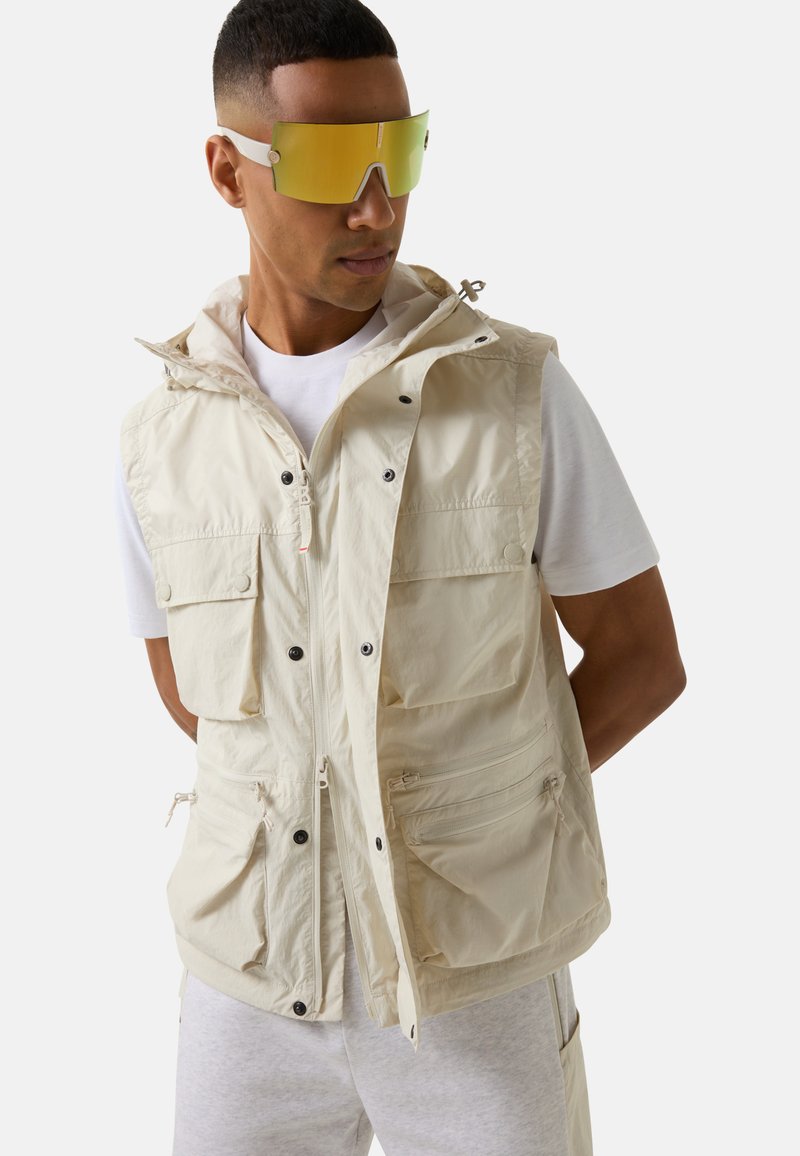 Beige ärmellose Weste mit mehreren Taschen, verstellbarem Kapuzenrand und Druckknöpfen vorne. Getragen über einem weißen T-Shirt und grauen Hosen.