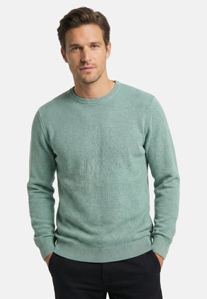 Mand med kort brunt hår iført en lysegrøn tekstureret langærmet sweater og sorte bukser, stående med hænderne i lommerne.