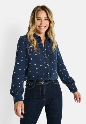 Chemise bleu marine à boutons avec broderie florale blanche, tissu texturé et manches bouffantes longues, assortie à un jean en denim foncé.
