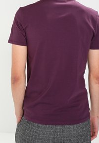 Camiseta de algodón de manga corta y ajustada en púrpura oscuro con un diseño de espalda liso. Cuello sin costuras y textura suave. Combinada con pantalones grises con estampado.