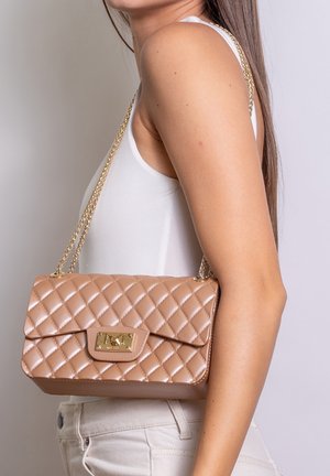 Donna con top bianco smanicato e pantaloni chiari, che porta una borsa beige trapuntata con tracolla catena dorata sulla spalla.