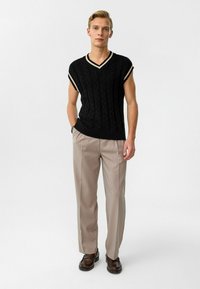 Zwarte gebreide mouwloze trui met V-hals en crèmekleurige details; gecombineerd met lichtbeige pantalon en donkerbruine schoenen. Model staat tegen een witte achtergrond.