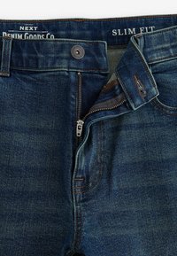 Jeans slim fit din denim de culoare albastru închis, prevăzute cu fermoar metalic și închidere cu buton, având un finisaj texturat subtil și cusături de contrast.