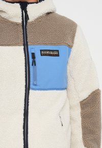Fleece jacket with a cream body, brown shoulder patches, and a blue chest panel. Features zip pockets and an embroidered brand logo.