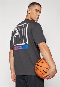 Tmavě šedé tričko s velkým bílým grafickým prvkem na zadní straně, zobrazující skákající postavu. Logo Reebok v mnoha barvách na spodní části. Basketbal v ruce.