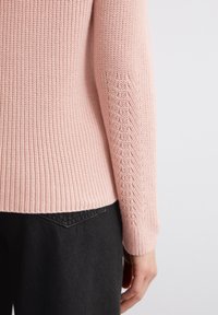 Hellrosa Strickpullover mit gerippter Struktur und Zickzackmuster an den Ärmeln, kombiniert mit schwarzen Jeans.