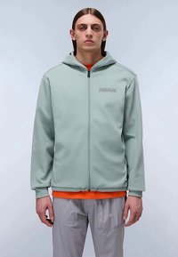 Ung mann med glatt brunt hår iført en lys grønn hettejakke, oransjefarget skjorte og lysegrå bukser, stående mot en enkel bakgrunn.