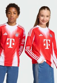 Maillot de football rouge à manches longues avec dégradé blanc, arborant les trois bandes Adidas, un logo de club et le logo du sponsor sur le devant.