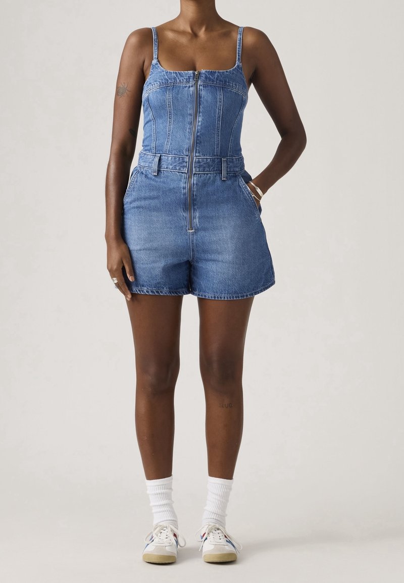 Femme portant une combinaison short en denim bleu avec fermeture éclair et fines bretelles, chaussettes blanches et baskets blanches, debout avec une main dans la poche sur fond uni.