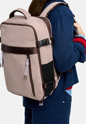 Osoba nosząca beżowy i brązowy plecak z wieloma suwakami i klamrami na ciemnoniebieskiej kurtce i spodniach, trzymająca pasek jedną ręką.