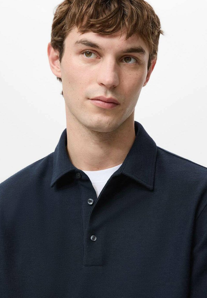 Polo bleu marine en tissu texturé, patte de boutonnage à trois boutons et col classique. Porté sur un sous-vêtement blanc, design minimaliste.