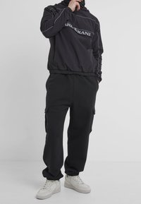 Person wearing black Karl Kani pullover, black cargo pants, and white sneakers, standing with one hand near the face and other in pocket.