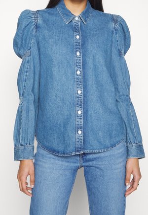 Chemise en denim bleu avec des manches bouffantes, fermeture à boutons et col classique. Texture lisse et design décontracté, associée à un jean bleu clair.