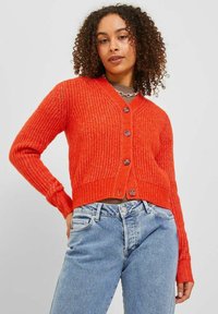 Cardigan rosso lavorato a maglia con scollatura a V, maniche lunghe e due bottoni frontali. Indossato sopra jeans azzurri a vita alta con texture a coste.