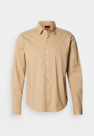 Camicia beige a maniche lunghe in cotone, con colletto button-down, bottoni frontali e un'etichetta rossa sul lato. Texture liscia.