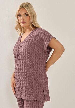 Lyshåret kvinde iført mauve tekstureret tunika og matchende bukser, stående med den ene hånd på hoften mod en ensfarvet lys baggrund.