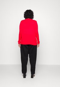 Persona con capelli ricci che indossa un top rosso a maniche lunghe, pantaloni neri larghi e scarpe nere con tacco, in piedi contro un muro bianco.