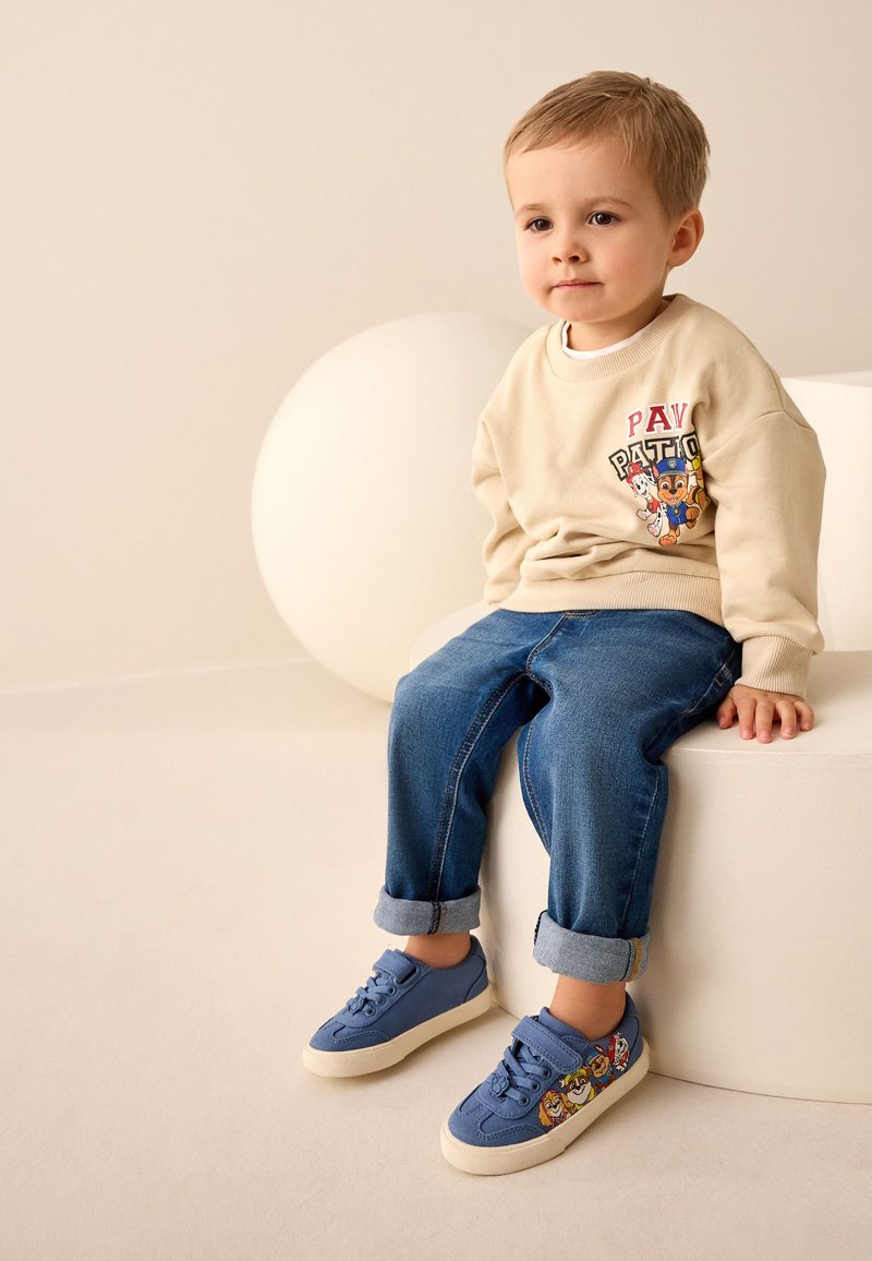 Sweat-shirt beige avec imprimé graphique, jean en denim bleu retroussé aux chevilles, et baskets bleues ornées de motifs de dessins animés colorés.