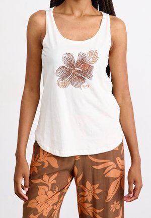 Femme portant un débardeur blanc à imprimé floral marron et un pantalon marron avec de grandes fleurs orange.