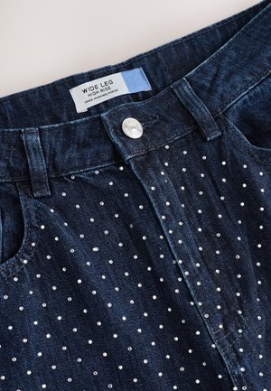 Ciemne dżinsy o szerokich nogawkach mają wysoki stan, ozdobione równomiernie rozmieszczonymi metalicznymi ćwiekami z przodu oraz srebrnym guzikiem do zapięcia.