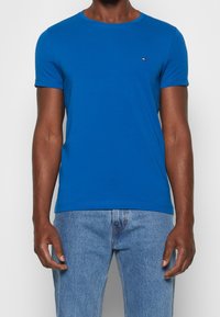 T-shirt en coton bleu à manches courtes avec un col rond, présentant un petit logo sur la poitrine et un design sans coutures. Associé à un jean en denim clair.