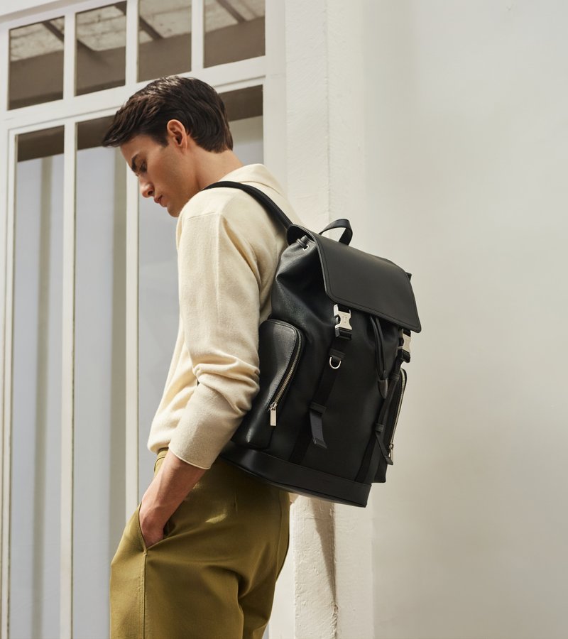 Junger Mann trägt beigen Pullover und olivgrüne Hose und lehnt an einer Wand mit einem schwarzen Lederrucksack auf dem Rücken.