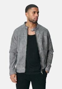 Veste en suédine grise avec col montant, fermeture éclair sur toute la longueur, poches latérales et texture lisse, associée à un t-shirt noir en dessous.