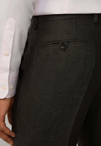 Pantaloni tailleur grigio scuro in tessuto testurizzato. Presentano una tasca sul retro con un solo bottone e sono abbinati a una camicia a righe rosa chiaro.