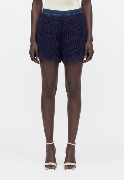 Le modèle porte un short en maille texturée bleu marine et des sandales à talons hauts blanches avec une bride à la cheville, debout devant un fond clair uni.