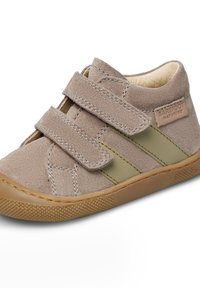 Scarpa in suede beige con due cinturini in velcro, dettagli verde oliva, punta arrotondata e suola in gomma testurizzata. Presenta un'etichetta di marca sul lato.