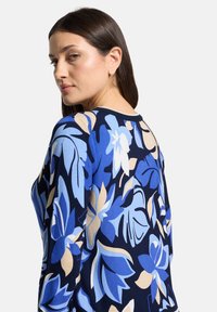 Femme aux cheveux bruns lisses portant un pull bleu marine avec de grands motifs floraux bleus, beiges et blancs, regardant par-dessus son épaule.
