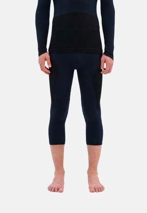 Man met een zwarte top met lange mouwen en cropped leggings, blootsvoets staand tegen een witte achtergrond.