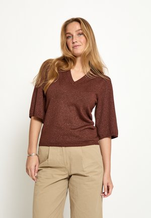 Vrouw met lang blond haar, gekleed in een bruin V-hals topje met korte mouwen en beige hoge taille broek, staand tegen een effen witte achtergrond.