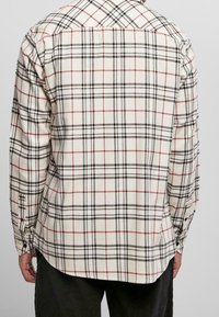 Man in een witte blouse met lange mouwen en een zwart-rood ruitpatroon, gezien vanaf de achterkant, gecombineerd met donkere broek.